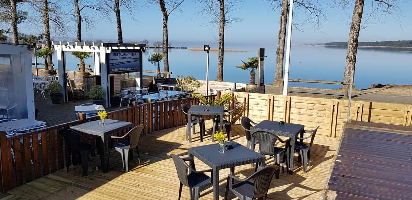 Terrasse en bois du Catamaran face au lac de Sanguinet, avec ses tables, parasols et palmiers